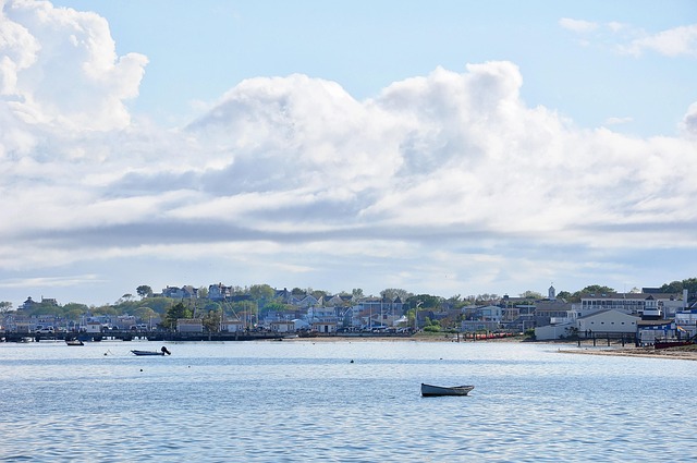 A quiet coastal harbor with small boats floating on calm water, shoreline buildings in the distance, and large white clouds filling a blue sky.