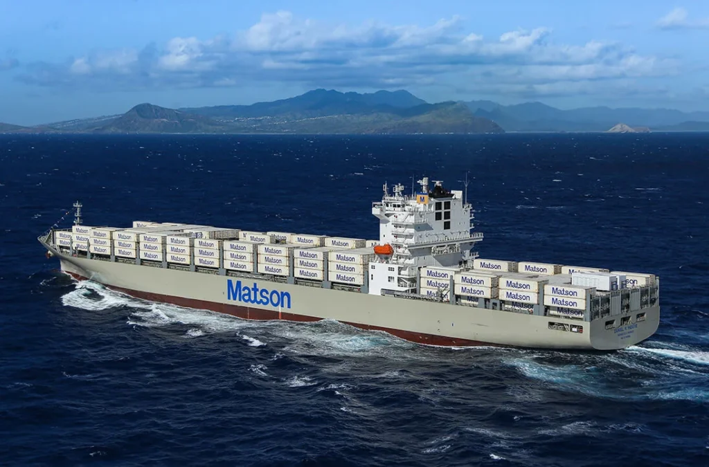 A Matson cargo ship loaded with shipping containers sails across deep blue ocean water with a mountainous island in the background under a partly cloudy sky.
