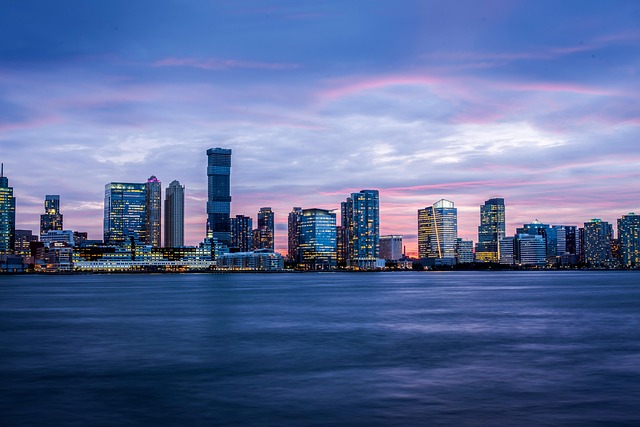 A city skyline at dusk with illuminated skyscrapers reflecting on calm water under a pink and purple evening sky.