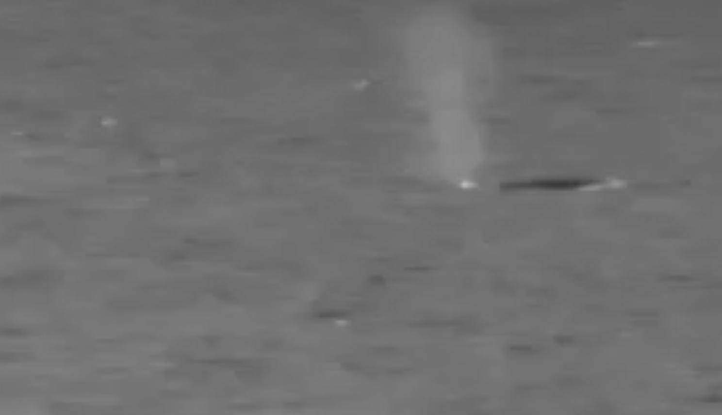 Black and white video still of a blue whale barely breaching and steam coming out of it's blowhole.